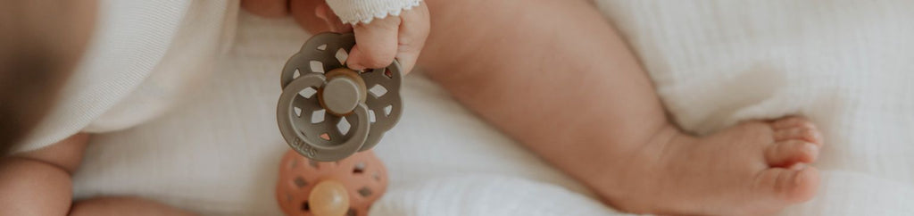 A baby holding a BIBS pacifier while lying down, illustrating a moment of play and comfort.