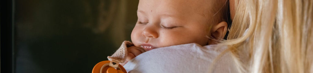 A baby peacefully sleeping on a caregiver's shoulder, holding a pacifier, illustrating the importance of a well-scheduled nap for optimal sleep.