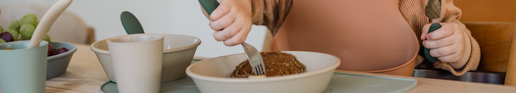 Toddler eating with a baby utensils, holding a fork and knife.