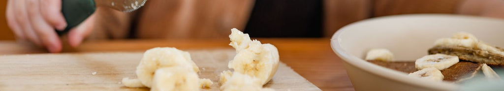 Toddler cutting banana slices with a baby utensils.